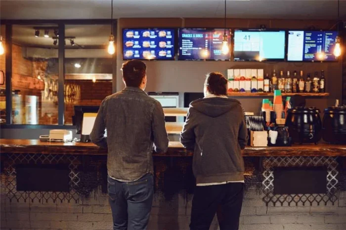 Two customers order at a fast casual restaurant with digital menu boards, highlighting a modern foodservice customer experience.