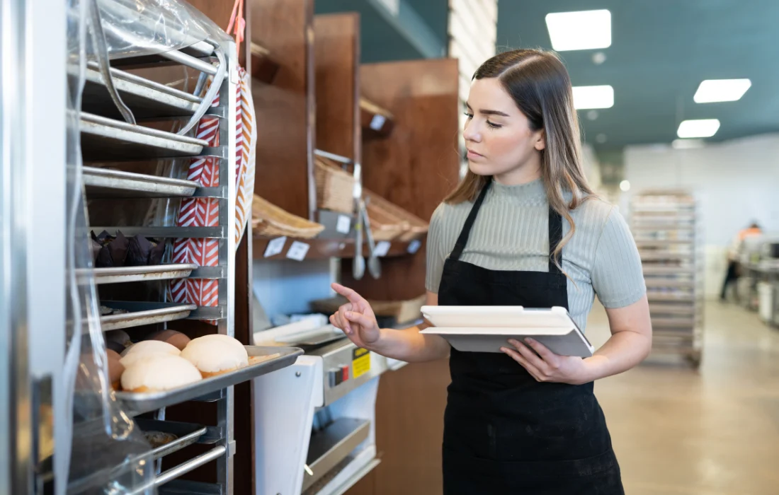 A woman in a bakery wearing an apron holds a tablet and inspects a tray of bread on a large industrial baking rack.