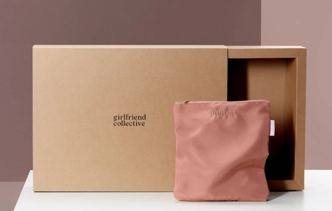 Large brown cardboard box with blush pink fabric pouch embroidered with brand logo placed in front.