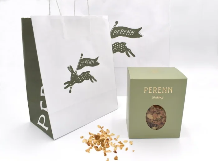 Perenn Bakery green granola box with window display next to branded white paper bag with hare logo and scattered granola.