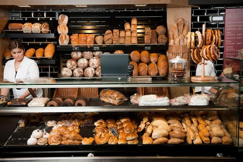 Bakery counter with loaves, rolls, and pastries on display, staff preparing orders behind the glass case.