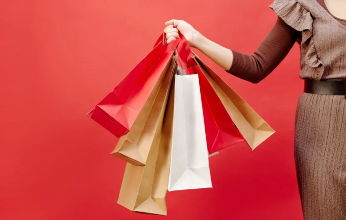 Person holding red, white, and brown paper shopping bags against a red background, representing retail packaging.