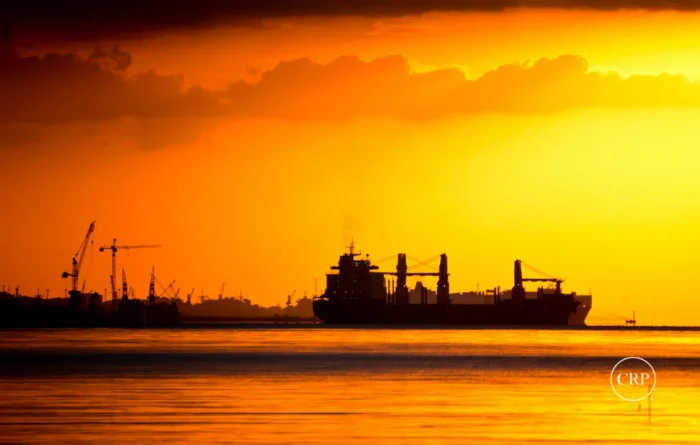 Cargo ship at sunset near port silhouettes reflects global trade disruption linked to rising gas prices and supply chain strain.